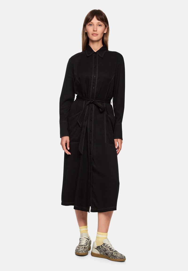 Schwarzes Hemdkleid mit langen Ärmeln, Knopfleiste vorne, Gürtel in der Taille und kontrastierenden Nähten. Kombiniert mit gemusterten Turnschuhen und gelben Socken.