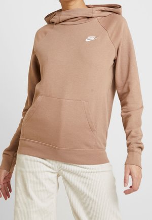 Beige Nike-hættetrøje med forlomme båret af en person iført hvide ribstrikkede bukser, vist fra nakken til midt på låret.