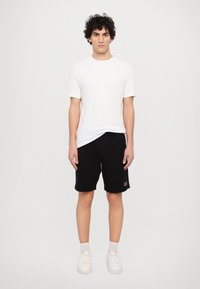 Hvid kortærmet t-shirt med et diskret logo, parret med sorte shorts med et logo i guld, suppleret af hvide sneakers.