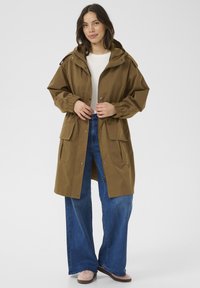 Bruine waterdichte parka met een capuchon, elastische manchetten, een trekkoord in de taille en twee voorkruiszakken, gecombineerd met wijde blauwe jeans en lichte schoenen.
