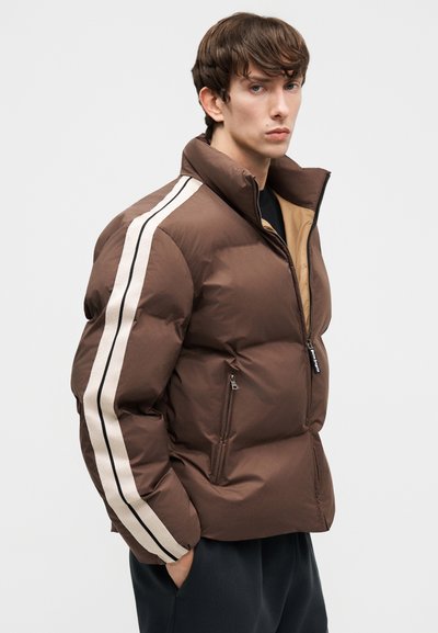 Junger Mann, der eine braune Steppjacke mit weißen und schwarzen Streifen an den Ärmeln trägt, steht mit einer Hand in der Tasche vor einem einfarbigen Hintergrund.