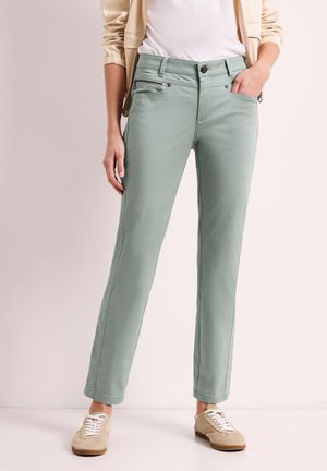 Kobieta ubrana w jasnozielone spodnie o prostych nogawkach, beżowe tenisówki, biały top i beżową kurtkę, stojąca na białej podłodze z ręką w kieszeni.