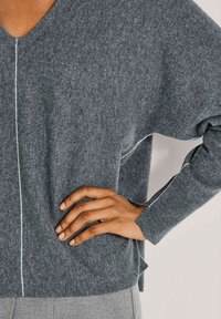 Person, der einen grauen Langarm-Strickpullover mit weißen Nahtdetails trägt und eine Hand auf die Hüfte legt.