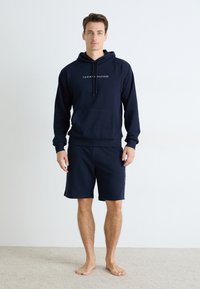 Sweat à capuche bleu marine avec poche avant et cordon de serrage, arborant le logo blanc "TOMMY HILFIGER". Associé à un short assorti, les deux en tissu doux.