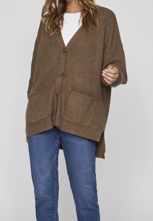 Bruine geribbelde cardigan met een v-hals, lange mouwen, twee voorzakken en een iets verlengde achterkant, gedragen over een witte top en blauwe jeans.