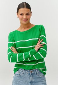 Grün gestreifter Pullover mit weißen horizontalen Linien, weicher Stoff, lockere Passform und weitem Ausschnitt. Kombiniert mit hochgeschnittenen Jeans.