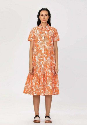 Femme portant une robe fleurie orange et blanche à manches courtes, longueur genoux, avec un col, debout les bras détendus le long du corps.