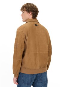 Brun suede bomberjakke med ribbet kant og mansjetter, featuring en tagdetalje på bagsiden og en sidelomme på ærmet.