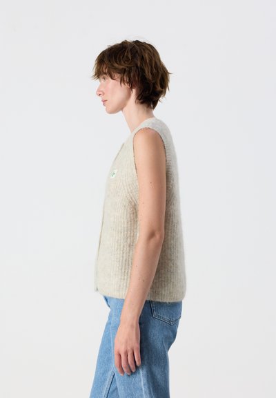 Femme aux cheveux bruns courts portant un gilet en tricot beige sans manches et un jean bleu, debout de profil sur un fond blanc uni.