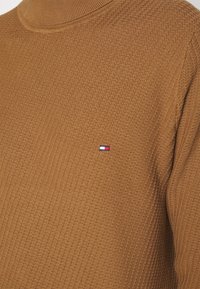 Maglione a collo alto marrone con un motivo testurizzato a costine. Presenta un piccolo logo ricamato in rosso, bianco e blu sul petto.