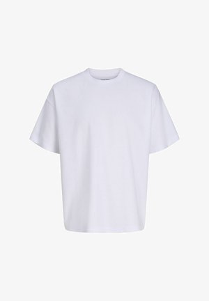 Jeune personne aux cheveux bouclés portant une chemise à carreaux bleue sur un t-shirt blanc, un jean foncé et des baskets noires, debout et souriante.