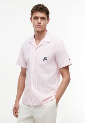 Młody mężczyzna ubrany w jasnoróżową i białą koszulę z krótkim rękawem w pionowe paski z kieszenią na piersi oraz białe spodnie.