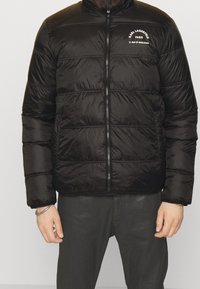 Svart puffjacka med dragkedja fram, trapetsmönster och vitt logotypdetalj på bröstet. Materialet verkar glansigt och lätt.