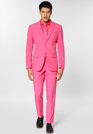 Lyserød jakkesæt med en tætsiddende blazer og matchende bukser. Glat stof, to forlommer, parret med et matchende slips og sorte sko.