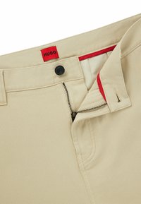 Beige trousers with a red interior waistband, featuring a zip fly and button closure. Textured fabric with visible stitching details.