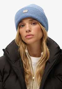Beanie lavorato a maglia di colore azzurro chiaro con texture a costine e logo a patch. Indossato con un giubbotto trapuntato nero e un maglione cremoso con zip.