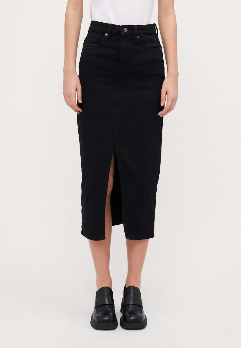 Mujer que lleva una falda midi negra de tiro alto con una abertura frontal y zapatos negros de slip-on, de pie contra un fondo claro y liso.