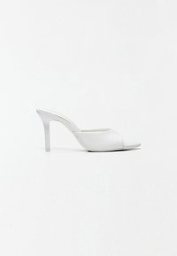 Mule blanche à talon haut avec bout ouvert, design à enfiler et talon stiletto étroit sur fond blanc uni.