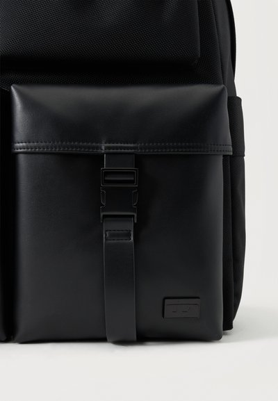 Poche avant de sac à dos noire avec un rabat sécurisé par une boucle en plastique noire et un petit logo rectangulaire en bas à droite.