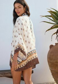 Kimono léger en creme avec des motifs floraux ; présente des rayures brunes et bleues à l'ourlet. Design ample et fluide avec des manches larges.