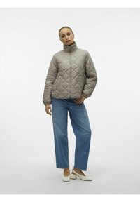 Gequilted, lichtgrijze jas met een hoge kraag en rits aan de voorkant, gecombineerd met wijde blauwe jeans en witte blokhakken.