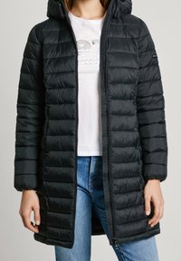 Svart puffjacka med en strukturerad quiltad design, huva och framdragkedja. Bärs över en vit skjorta och blå jeans.