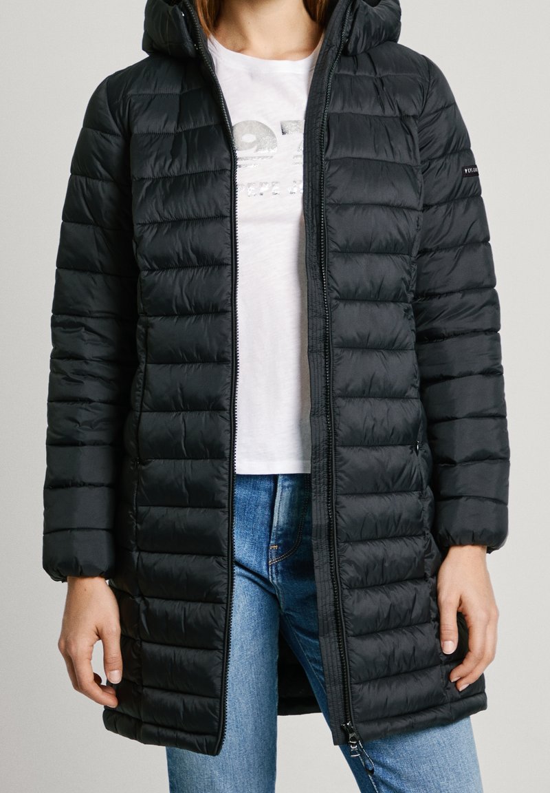 Svart puffjacka med en strukturerad quiltad design, huva och framdragkedja. Bärs över en vit skjorta och blå jeans.