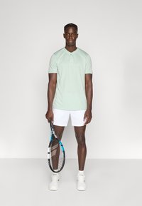 Ανοιχτοπράσινη πόλο μπλούζα με πατιλέτα τριών κουμπιών, σε συνδυασμό με λευκά σορτς, κρατώντας ρακέτα τένις. Λείο ύφασμα, αθλητική εφαρμογή.