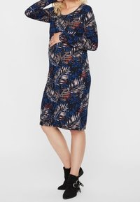 Langärmliges Umstandskleid mit navyblauem Grund und buntem Blumenmuster, figurbetonter Passform und weichem Stoff. Kombiniert mit Stiefeletten.
