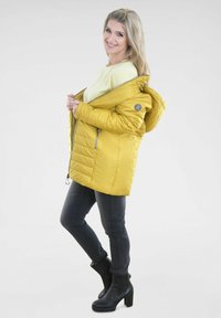Gelbe, gesteppte Daunenjacke mit Kapuze, Reißverschlusstaschen und Logodetail, kombiniert mit schwarzen Jeans und schwarzen Stiefeln mit Absatz.