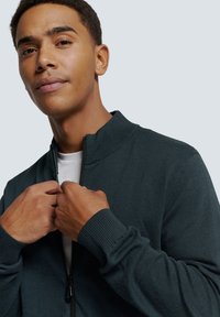 Donkergroene zip-up trui met hoge kraag, ribbelmanchetten en een gladde textuur, gedragen over een wit t-shirt, handen die de voorkant aanpassen.