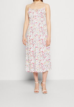 Kvinde iført en hvid ærmeløs midi-kjole med et pink og orange blomster mønster samt hvide hæl-sandaler, stående mod en ensfarvet baggrund.