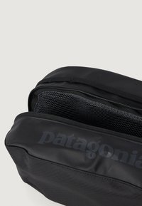 Crna torbica na zatvarač izrađena od izdržljivog materijala s mrežastim interijerom. Ima logo "Patagonia" sa strane i glatku teksturu.