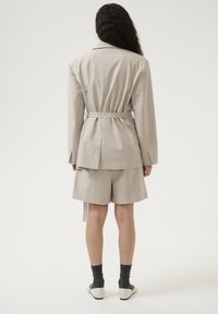 Beige karierter Blazer mit einem taillierten Gürtel, kombiniert mit passenden Shorts. Glatter Stoff, taillierte Passform und lange Ärmel.