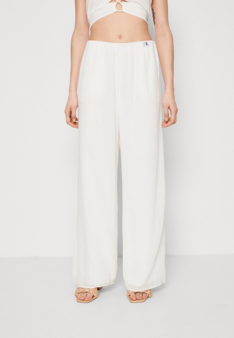Femme portant un pantalon blanc taille haute à jambes larges avec une petite étiquette logo Calvin Klein, associé à des sandales tissées beige.