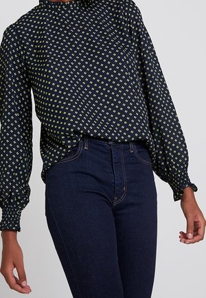 Person, die eine marineblaue Bluse mit langen Ärmeln und kleinem gelbem Kariermuster sowie hochtaillierte dunkelblaue Jeans trägt, steht vor einem einfarbigen Hintergrund.