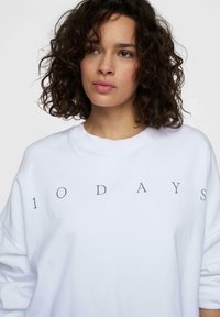 Mujer joven con cabello rizado y oscuro que lleva una sudadera blanca con el texto "10DAYS" impreso en el pecho.
