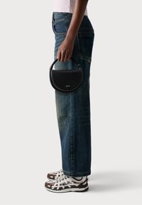 Sac à main en cuir noir de forme semi-circulaire, avec des accents métalliques et une poignée supérieure, associé à un jean large délavé bleu et des baskets marron.