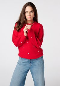 Pull rouge avec motif de cœur, manches longues et encolure arrondie, associé à un jean bleu. Le tissu semble doux et texturé.