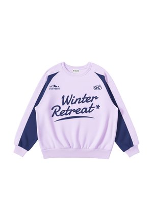 Young man with curly hair wears a purple and navy sweatshirt with "Winter Retreat" and mountain logo, hands in pocket, neutral background.