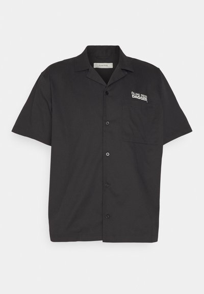 Īsas piedurknes melna krekls, kas izgatavots no gludas auduma, ar krūšu kabatu un baltu logo kreisajā pusē. Pogas aizdare priekšpusē.