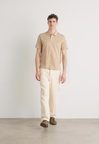 Chemise polo beige avec un col court, associée à un pantalon couleur crème. Modèle portant des chaussures vert olive, se tenant devant un fond neutre.