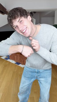 Šedý žebrovaný svetr, stříbrný řetízkový náhrdelník a náramek, světle modré džíny. V pozadí dřevěná podlaha a hnědá pohovka.