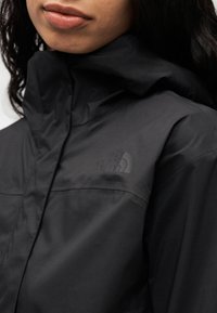 Μαύρο αδιάβροχο μπουφάν με ψηλό γιακά, που διαθέτει διακριτικό λογότυπο στο στήθος. Το ύφασμα έχει ομαλή υφή και κομψό σχεδιασμό.