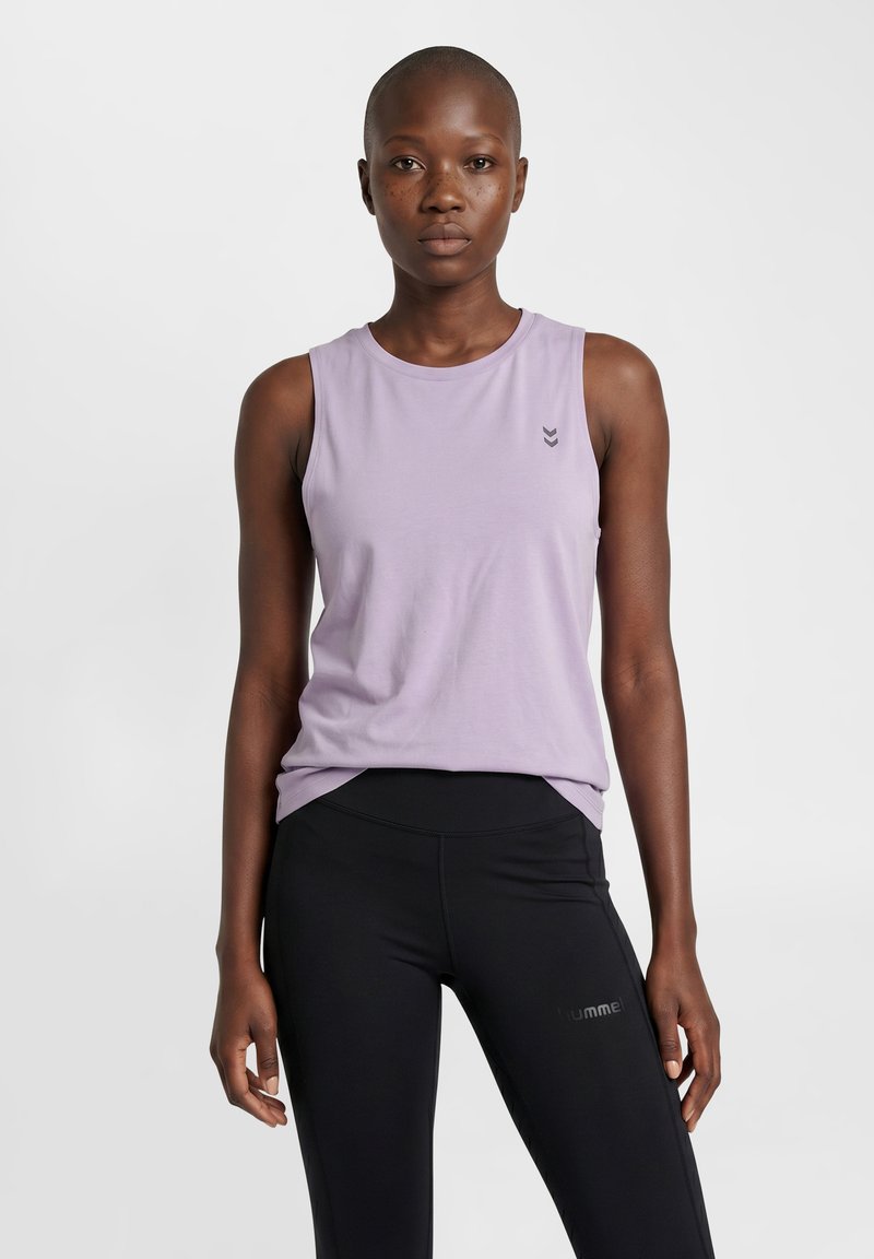 Woman with short hair wearing a sleeveless light purple top and black athletic leggings standing against a plain light background.