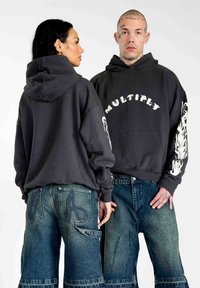 Grå hættetrøje med "MULTIPLY" i hvid tekst og flammer på ærmerne; parret med brede blå denimbukser med dekorativ syning.