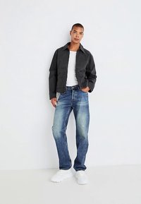 Crna jakna s ovratnikom, bijela majica, svijetloplave traperice i bijele tenisice. Model stoji na jednostavnoj pozadini.