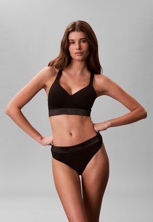 Femme aux longs cheveux bruns portant un soutien-gorge Calvin Klein noir et une culotte assortie, debout les mains sur les hanches devant un fond gris uni.