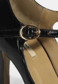 Schwarze lacklederne High Heels mit einem schlanken Riemen, der eine goldene ovale Schnalle trägt. Glatte Textur und beiges Fußbett sichtbar.