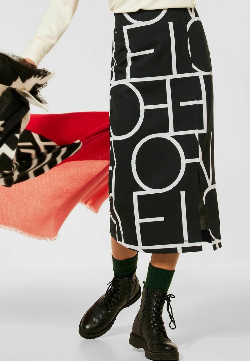 Jupe noire ornée de grandes lettres graphiques blanches. Longueur genou avec une coupe droite et une fente latérale, portée avec des bottes de combat noires et des chaussettes vertes.